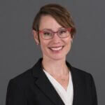 Ashley is smiling and has short brown hair and is wearing glasses and a black jacket