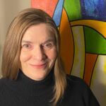 Barbara has long blonde hair and is wearing a black turtleneck in front of a multicolored mural