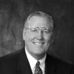 Fred has short gray hair and glasses and is smiling and wearing a dark suit and tie