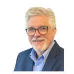 Gene is wearing a navy suit jacket over a blue button-up shirt.