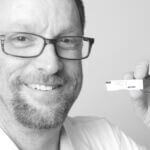 John has a goatee and glasses and is smiling and holding a piece of paper that says access