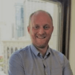 Jonathan has a beard and is wearing a blue button-down shirt, standing in front of a window.