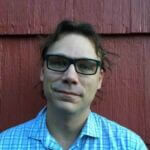 Joshua has short brown hair and has glasses wearing a blue checked shirt