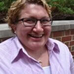 Katie is smiliing and wears glasses and a pink shirt