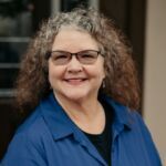 Kristi is smiling and has curly brown hair and wears glasses and a blue shirt.