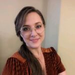 Leah has long brown hair and glasses and is smiling