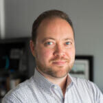 Mark has short dark hair and a beard and mustache and is wearing a light blue checked shirt.