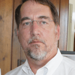 Mark has a gray goatee and is wearing glasses and he is wearing a white shirt