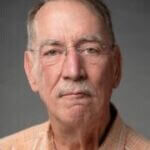 Headshot of Mark Mazz, a man with short gray hair, a gray mustache, and rimless eyeglasses, wearing an orange and white plaid collared shirt against a neutral gray background.