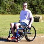 Mick is smiling and sitting in a wheelchair in front of a hay bale