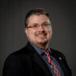 Randy is smiling and wearing glasses and a dark suit.