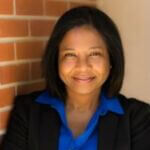 Carol is grinning and has medium length black hair and is wearing a blue shirt and black jacket