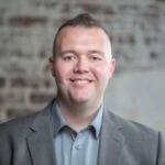 Robert is smiling and wearing a gray suit and standing in front of a brick wall