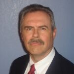 Robert has short gray hair and a mustache and is wearing a dark suit and red tie