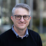 Steve has short hair and is wearing glasses and a dark sweater