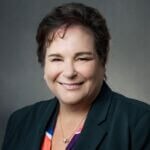 Terry has short brown hair and is wearing a dark jacket and bright top and is smiling